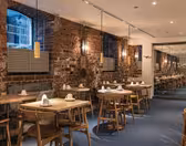 Et hyggeligt restaurantinteriør med synlige murstensvægge og varm belysning. Træborde og stole er pænt arrangeret, hvert bord dækket med hvide servietter. Store spejle og vinduer øger rummeligheden, mens pendellamper giver stemningsbelysning.