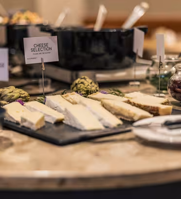 En række forskellige oste er udstillet på et rundt marmorbord med et mærket skilt med "Ostevalg". I nærheden er der krukker, artiskokker og redskaber. Indstillingen er indendørs, sandsynligvis ved en buffet eller spiseplads.