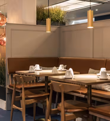 Et hyggeligt restaurantinteriør med træborde og -stole, sat til spisning med hvidt service. Brune læderbænke langs væggene, dekoreret med planter. Varme lysarmaturer hænger fra loftet og skaber en indbydende atmosfære.