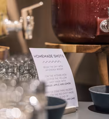 Drikkeautomater i glas med tud sidder på træstandere, den ene fyldt med en gul væske og den anden med rød. Et skilt med titlen "Homemade Shots" er synligt, omgivet af små glaskopper og to blå skåle.