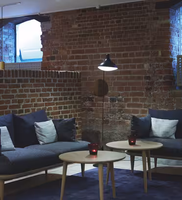 Et hyggeligt værelse med synlige murstensvægge har to moderne sofaer med hynder, et sofabord i træ og en høj gulvlampe. Blød belysning og store vinduespartier skaber en indbydende atmosfære.