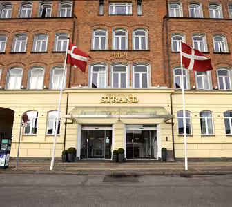 Billedet viser indgangen til Copenhagen Strand Hotel, en bygning med gul facade og røde murstens øverste etager. To danske flag er vist foran. Over indgangen står "1869" på murværket.