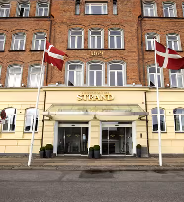 Billedet viser indgangen til Copenhagen Strand Hotel, en bygning med gul facade og røde murstens øverste etager. To danske flag er vist foran. Over indgangen står "1869" på murværket.