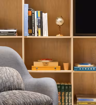 En hyggelig læsekrog med en moderne grå lænestol foran en træhylde. Hylderne rummer bøger, en globuspynt, en trækasse og et Scrabble-spil. En del af et fladskærms-tv er synligt til højre.