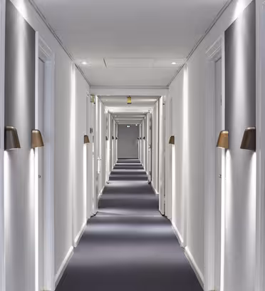 En lang, veloplyst hotelgang med blåt tæppe og hvide vægge. Døre med rumnumre og oplyste væglamper er jævnt fordelt på begge sider, hvilket fører til et fjernt udgangsskilt for enden af korridoren.