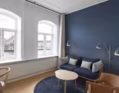 Moderne stue med et stort buet vindue, blå accentvæg og trægulv. Har en blå sofa med hynder, læderstol, rundt træbord og gulvlamper. Uden for vinduet ses en naturskøn udsigt over en flod og bygninger.