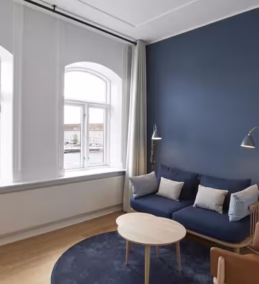 Moderne stue med et stort buet vindue, blå accentvæg og trægulv. Har en blå sofa med hynder, læderstol, rundt træbord og gulvlamper. Uden for vinduet ses en naturskøn udsigt over en flod og bygninger.