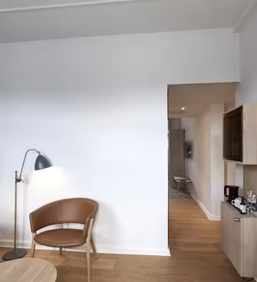 Moderne hotelværelse med en blå accentvæg, trægulve og elegante møbler. Den har et rundt træbord, en brun stol, en blå sofa med puder, stående lamper, et væghængt tv og en lille åben reol med kaffemaskine.