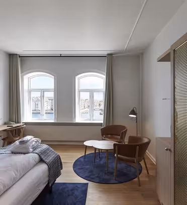 Et moderne hotelværelse med en dobbeltseng, et trægarderobeskab og et rundt sofabord med to læderstole. Rummet har store vinduespartier med lysgardiner, et væghængt tv, samt udsigt til vand og bygninger udenfor.