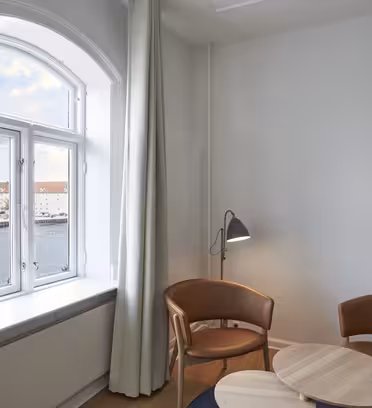 Et minimalistisk værelse med to brune stole og et lille rundt træbord. Et stort vindue afslører en udsigt over vandet. En høj gulvlampe er placeret ved siden af stolene, og et lysgardin dækker delvist vinduet.