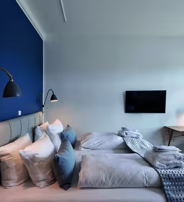 Et moderne hotelværelse med to pænt lavet enkeltsenge mod en blå accentvæg. Et væghængt tv er overfor sengene. Et lille skrivebord med en lampe er nær et vindue med lukkede gardiner. Rummet er lyst og minimalistisk i stilen.