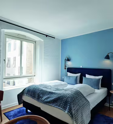 Et moderne hotelværelse med en stor seng dækket af blåt og hvidt sengetøj. Der er en lyseblå accentvæg, to vægmonterede lamper, et vindue med beige gardiner, et træskrivebord, en stol og et tv. Rummet har et minimalistisk, men hyggeligt design.