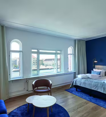 Et moderne hotelværelse med en stor seng, blå accentvæg og lyst trægulv. Der er et rundt bord med stole, en marineblå sofa og høje vinduer med udsigt over byen. Blød belysning skaber en hyggelig atmosfære.
