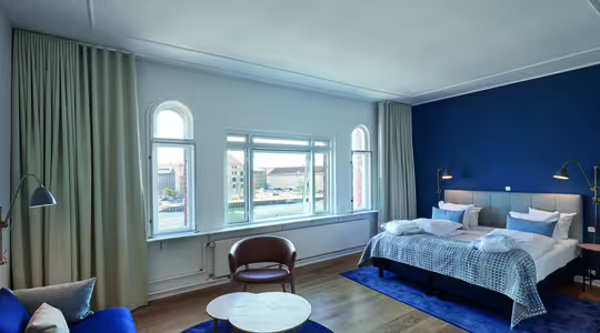 Et moderne hotelværelse med en stor seng, blå accentvæg og lyst trægulv. Der er et rundt bord med stole, en marineblå sofa og høje vinduer med udsigt over byen. Blød belysning skaber en hyggelig atmosfære.