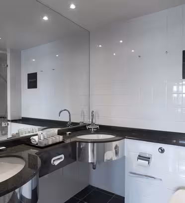 Et moderne badeværelse med to håndvaske og en sort bordplade. Væggen er flisebelagt hvid, og der er spejle over vaskene. Toiletartikler er pænt arrangeret på disken. En toilet- og toiletpapirholder er synlig til højre.
