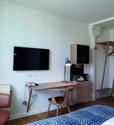 Minimalistisk hotelværelse med vægmonteret tv over et træskrivebord, udstyret med lampe og telefon. En hyggelig stol sidder i nærheden. En reol rummer kaffefaciliteter. Sengen har et mønstret blåt betræk, og en slank væglampe giver lys.