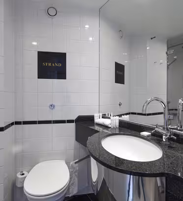 Moderne badeværelse med hvide flisevægge og mørk bordplade. Indeholder en rund håndvask, rustfrit stålhane, toilet og glasindkapslet bruser. Et spejl reflekterer rummet, og "Strand" er skrevet på to fliser.