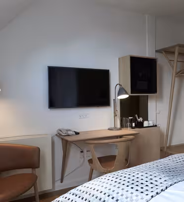 Moderne hotelværelsesinteriør med et fladskærms-tv monteret på en hvid væg, et træskrivebord med lampe og telefon, en stol med brunt betræk og et åbent klædeskab i træ med bøjler. Sengen er synlig med et hvidt og gråt tæppe.