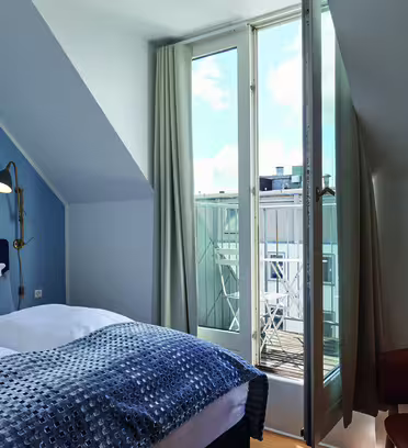 Et hyggeligt soveværelse har en seng med blåt og hvidt sengetøj, en gulvlampe og en lænestol. Store glasdøre åbner ud til en lille balkon med udsigt til nabobygningerne. Væggene er lyseblå og supplerer de rolige, luftige omgivelser.