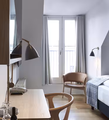 Et moderne hotelværelse med en seng, et skrivebord med en stol og en væghængt lampe. To store vinduer er beklædt med lysgardiner. En træstol og et lille sidebord står i hjørnet, hvilket skaber en hyggelig atmosfære.