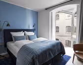 Et moderne hotelværelse med en blå accentvæg har en dobbeltseng med hvidt og blåt sengetøj. To væghængte læselamper flankerer sengen. Et stort vindue med beige gardiner giver udsigt til en tilstødende murstensbygning. En brun stol er placeret i nærheden.