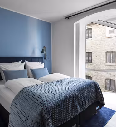 Et moderne hotelværelse med en blå accentvæg har en dobbeltseng med hvidt og blåt sengetøj. To væghængte læselamper flankerer sengen. Et stort vindue med beige gardiner giver udsigt til en tilstødende murstensbygning. En brun stol er placeret i nærheden.