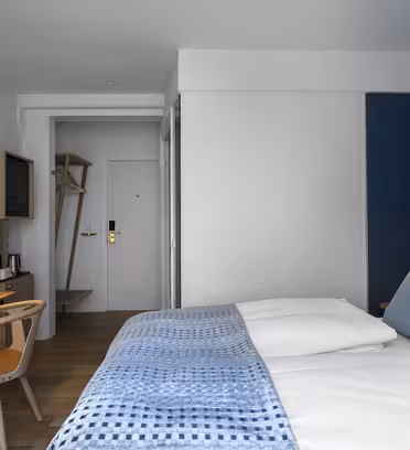 Moderne hotelværelse med en pæn opredt seng med hvidt og blåt sengetøj. Et træbord med stol og lampe er placeret op ad væggen. I baggrunden er der et minimalistisk åbent skab og indgangsdøren. Værelset har en hyggelig, rolig atmosfære.