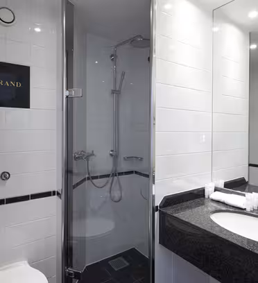 Moderne badeværelse med hvide klinker, lukket glas bruseområde og sort bordplade med håndvask. Et lille hvidt håndklæde og toiletartikler står på disken. Ordet "STRAND" er på flisen ved siden af bruseren.