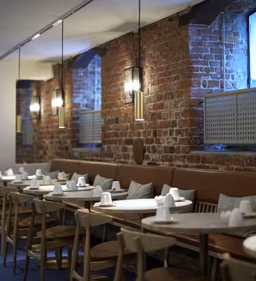 Et moderne restaurantinteriør med synlige murstensvægge og rækker af træborde sat med hvidt service. Hængende pendellamper oplyser rummet, og der er brune læderbænke langs væggen med puder til siddepladser.