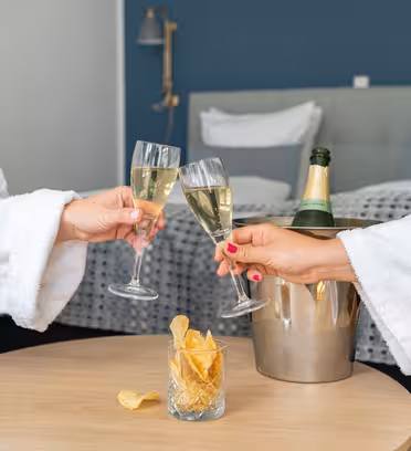 To personer i hvide rober skåler med champagneglas på et stilfuldt hotelværelse. En flaske champagne hviler i en isspand på bordet sammen med et glas fyldt med kartoffelchips. Plys sengetøj er synligt i baggrunden.