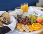 En morgenmadsbakke med rundstykker i en kurv, tre glas yoghurt toppet med müsli, to glas appelsinjuice og en tallerken med diverse frugter, herunder vindruer, æbler, appelsinskiver og cantaloupeskiver.