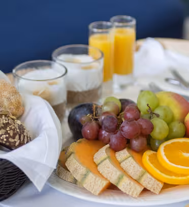 En morgenmadsbakke med rundstykker i en kurv, tre glas yoghurt toppet med müsli, to glas appelsinjuice og en tallerken med diverse frugter, herunder vindruer, æbler, appelsinskiver og cantaloupeskiver.