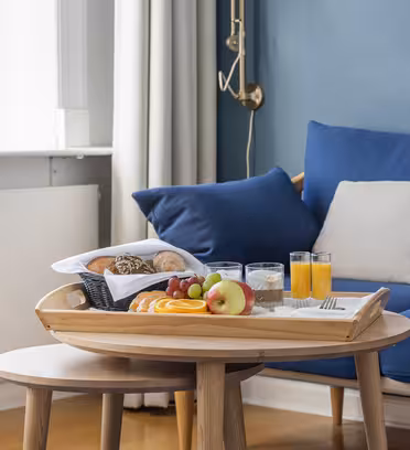 En træbakke på et sofabord viser en morgenmad med rundstykker, frugtskiver, druer, et æble, glas vand og appelsinjuice. Det ligger i en hyggelig stue med en blå sofa og beige puder nær et vindue.