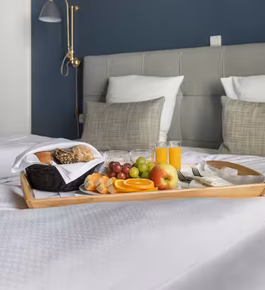 Morgenmadsbakke på en seng med to hvide dyner og en grå sengegavl. Bakken rummer juice, diverse frugter, rundstykker og en croissant. Der er en væghængt lampe over sengen, og værelsesvæggen er malet blå.