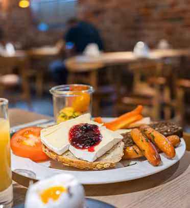 En tallerken med morgenmadsmad inklusive brød med ost og marmelade, tomatskiver, gulerødder og pickles står på bordet. Et glas juice, et glas mælk og et blødkogt æg er i nærheden. Baggrunden byder på en hyggelig café med murstensvægge og træstole.