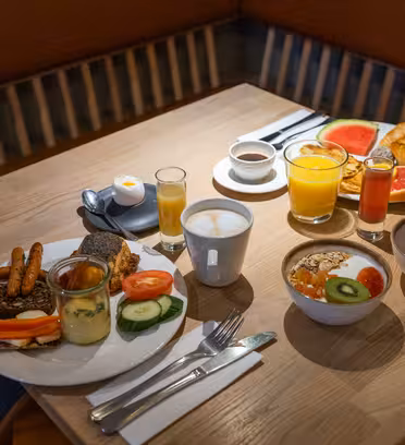 Et morgenbord med forskellige fødevarer, inklusive en tallerken med brød, kringler, ost og grøntsager, en cappuccino, juice og en skål kornprodukter med frugt. En anden tallerken rummer kager, vandmelon og et kogt æg i en æggekop.